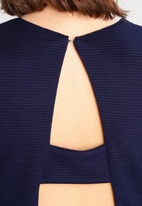 Námořnický žebrovaný top s zaobleným otvorem na zádech a malým zapínáním na knoflík. Design zahrnuje přiléhavou vnitřní bandu pod výřezem.