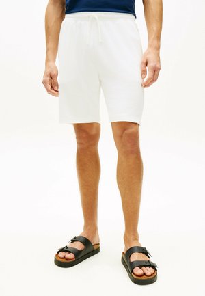 Mężczyzna w prostych białych szortach z troczkami i czarnych sandałach z klamrą, stojący na białym tle.