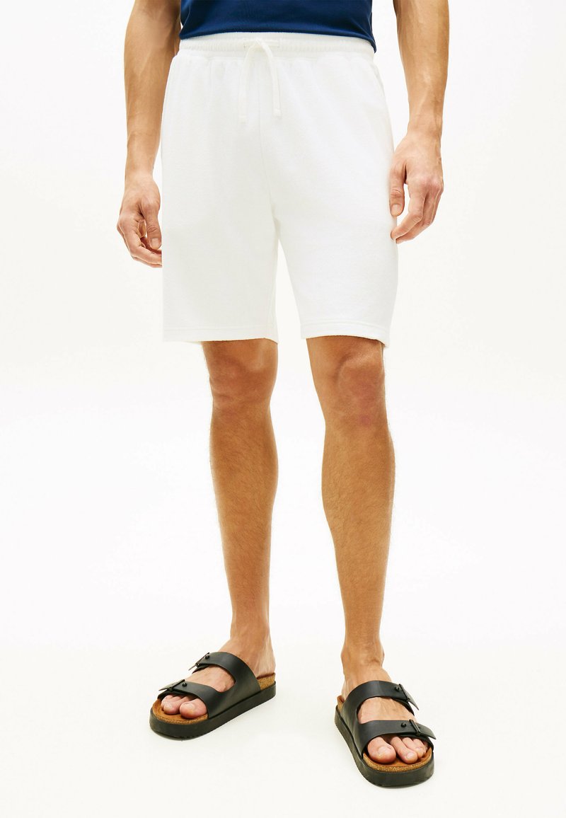 Mann trägt schlichte weiße Shorts mit Kordelzug und schwarze Sandalen mit Schnalle, steht vor einem weißen Hintergrund.