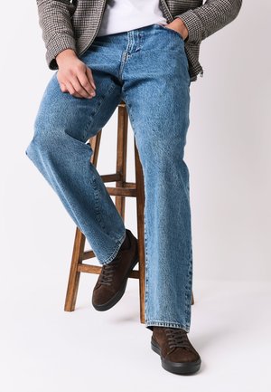 Persona con vaqueros azules, zapatillas marrones y chaqueta de cuadros sentada en un taburete de madera contra un fondo liso.