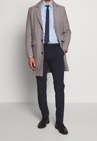 Grijze jas over een lichtblauw shirt, marineblauwe stropdas en donkere broek. Zwarte schoenen complementeren de outfit. De jas heeft twee voorzakken en knopen.