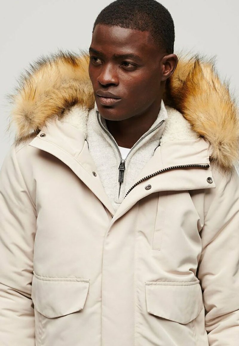 Beige Parka-Jacke mit gefütterter Kapuze, Reißverschluss, zwei Fronttaschen und einem weichen grauen Innenfutter. Glatte Textur, lässiges Design.