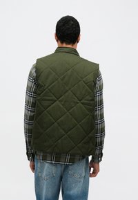 Πράσινο γιλέκο με καπιτονέ σχέδιο και γιακά, φορεμένο πάνω από ένα γκρι και μαύρο πουκάμισο με καρό. Το ύφασμα έχει λεία, υφή με υφαντό φινίρισμα.