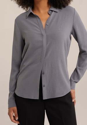 Kobieta ubrana w szarą koszulę na guziki z długim rękawem i czarne spodnie, stojąca z jedną ręką na biodrze na neutralnym tle.