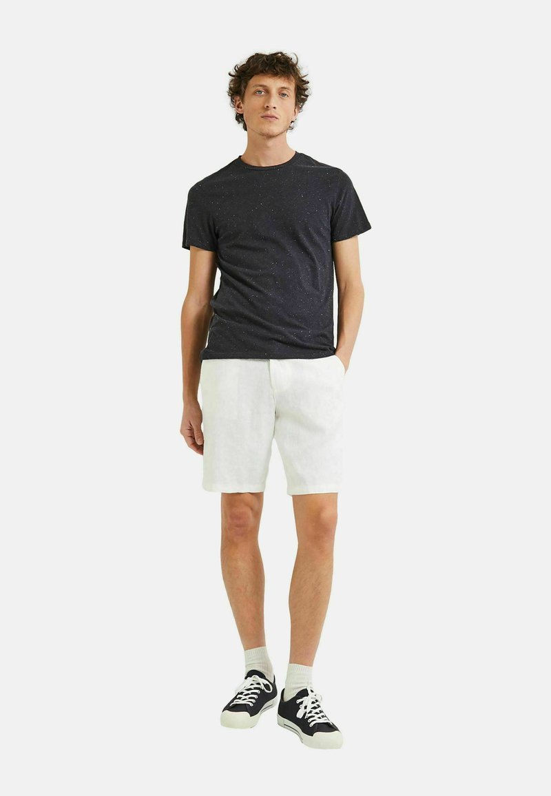T-shirt noir à motifs tachetés à manches courtes, associé à un short en lin blanc et des baskets noires avec des semelles et des lacets blancs. Tenue décontractée et légère.