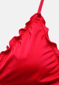 Haut de bikini en satin rouge avec un bord festonné et des bretelles ajustables. Texture lisse avec un détail de coutures froncées sur un côté.