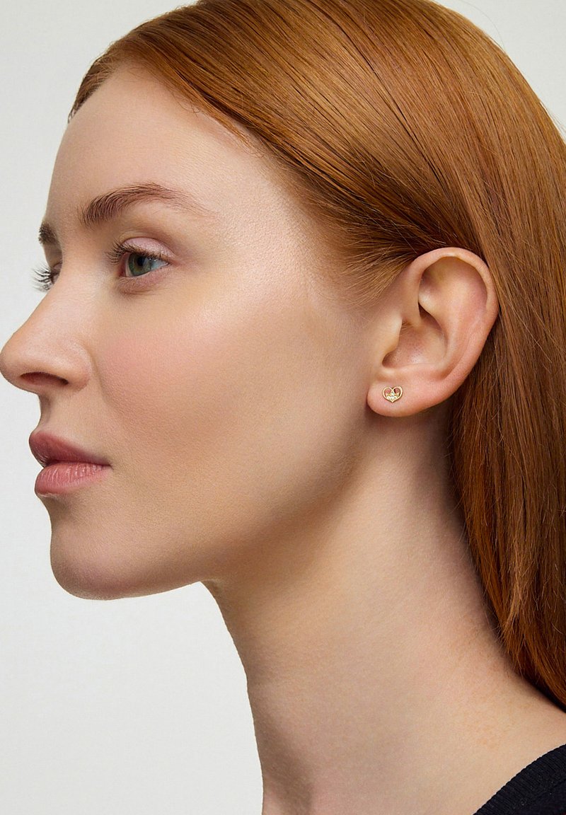 Złote kolczyki w kształcie serca na lewym uchu modelki; gładka metalowa powierzchnia z małym akcentem w postaci kamienia szlachetnego. Modelka ma proste czerwone włosy.