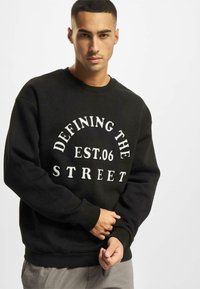 Mustapaita, jossa on valkoista tekstiä, joka lukee "DEFINING THE STREET EST.06". Ribbaatut kaulus ja hihat, rento istuvuus.