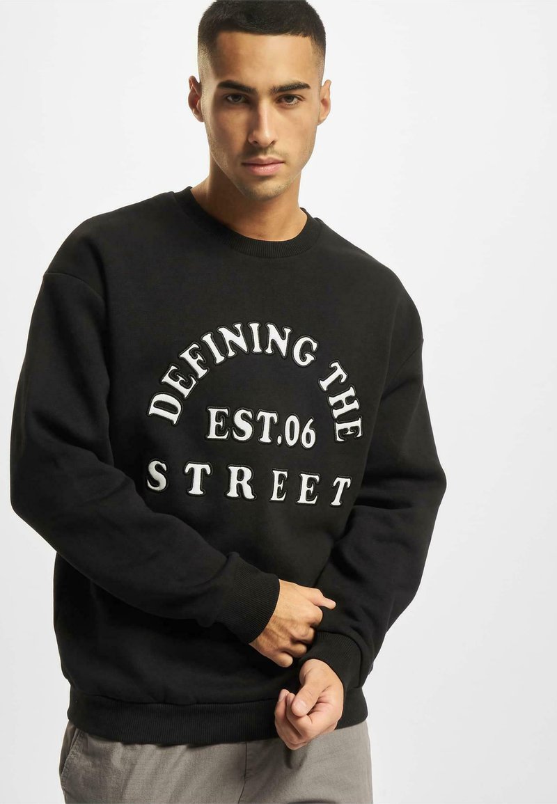 Mustapaita, jossa on valkoista tekstiä, joka lukee "DEFINING THE STREET EST.06". Ribbaatut kaulus ja hihat, rento istuvuus.