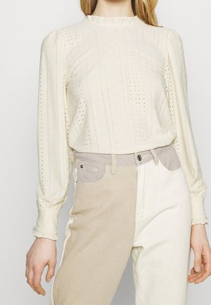 Vrouw draagt een crèmekleurige blouse met lange mouwen met eyelet-patronen en tweekleurige beige en witte broek met hoge taille.