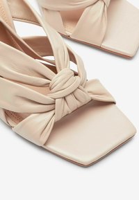 Sandales en cuir beige à bouts carrés, avec un détail de nœud torsadé sur les brides. Texture lisse avec un éclat subtil et un design minimaliste.