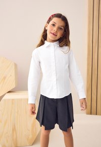 Chemise blanche à manches longues avec boutons, associée à un short plissé gris. La chemise présente un col classique et un design épuré, mettant en valeur une texture lisse.