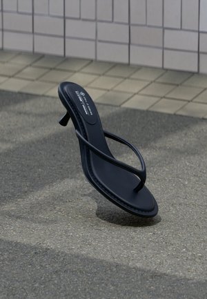 Μαύρο σανδάλι με λουράκια και τακούνι που αιωρείται πάνω από υφή γκρι πεζοδρομίου με φόντο τοίχο με πλακάκια.