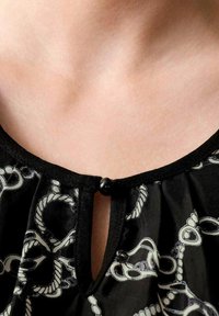 Schwarz-bluse mit einem Schlitz-Ausschnitt, der einen kleinen schwarzen Knopf und weiße, seilartige Muster aufweist. Semitransparente Stoff mit glatter Textur.