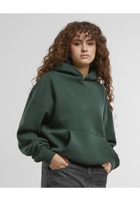 Overdimensjonert grønn hettegenser laget av mykt stoff, med et frontlomme og ribbede mansjetter. Enkelt design, ingen synlige mønstre eller detaljer.