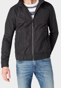 Giacca nera con zip e colletto alto, dotata di due tasche frontali e orlo elasticizzato. Indossata con jeans blu e una camicia a righe sotto.