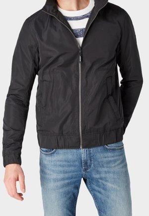 Veste zippée noire avec col montant, comportant deux poches avant et un ourlet élastiqué. Porte avec un jean bleu et une chemise rayée en dessous.