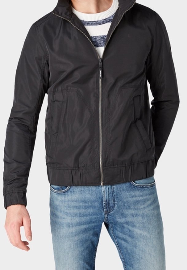Giacca nera con zip e colletto alto, dotata di due tasche frontali e orlo elasticizzato. Indossata con jeans blu e una camicia a righe sotto.