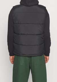 Svart pufferväst med horisontella sömmar, lagerdesign och en slät textur. Bärs över en svart hoodie med gröna byxor synliga nedanför.