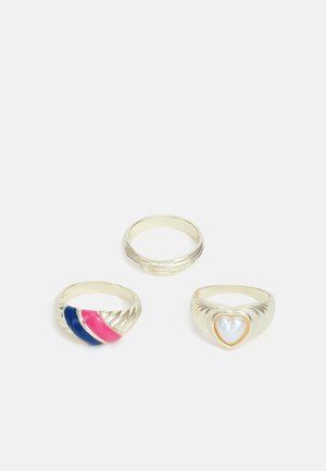 Três anéis: um com bandas de esmalte rosa e azul, um anel de prata liso e um anel em forma de coração com acabamento em pérola e contorno dourado.