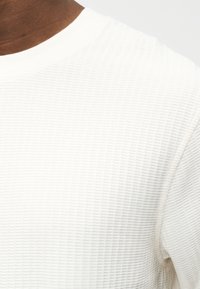 Κοντινό πλάνο ενός ατόμου που φοράει λευκό, υφασμάτινο κοντομάνικο πουκάμισο με στρογγυλή λαιμόκοψη, σε απλό φωτεινό φόντο.