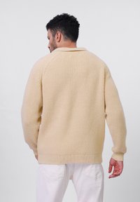 Beige stickad tröja med ribbat mönster, med en avslappnad passform och rundad krage, bärs över vita byxor, visad bakifrån.