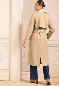 Beige trenssi takki, jossa on vyötärölle solmittava vyö ja liehuva takakuvio. Yhdistetty liehuviin farkkuihin ja vaaleisiin korkokenkiin.