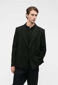 Μαύρο διπλό πλέγμα σακάκι με μεγάλους κουμπιά, δομημένους ώμους και λείο υλικό, φορεμένο πάνω από μαύρο πόλο μπλουζάκι.