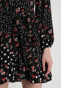 Abito nero con pois bianchi e motivi floreali rossi, caratterizzato da vita annodata, maniche lunghe e orlo arricciato. Tessuto dalla texture morbida.