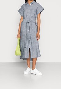 Robe chemise rayée en bleu et blanc, dotée d'une ceinture, de manches courtes, d'une fermeture à boutons et d'une fente jusqu'aux genoux, associée à un sac vert et des baskets blanches.