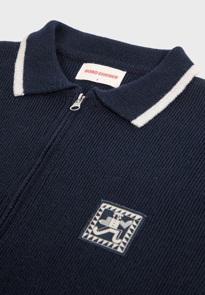 Marinblå stickad jacka med vitrandig krage, dragkedja fram och en logopatch med en geometrisk design på bröstet.