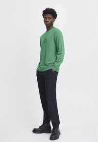 Maglietta verde a maniche lunghe con scollo rotondo e dettaglio del logo. Abbinata a pantaloni scuri e stivali neri con lacci. Design semplice e dalla vestibilità rilassata.