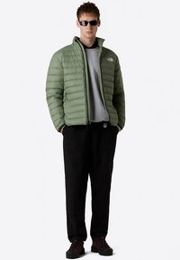 Gewatteerd groen jack met rits aan de voorkant en lange mouwen, gedragen over een grijs shirt en zwarte broek, gecombineerd met donkere schoenen en een zonnebril.