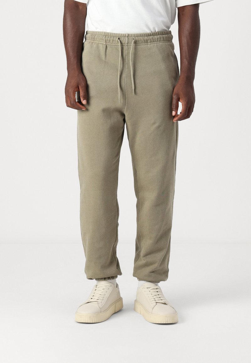 Grågrønne joggebukser med elastisk midje og snor, med innsnevrende ben og ribbestrikkede mansjetter, sammen med lyse sneakers.