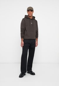 Felpa con cappuccio marrone e tasca frontale, abbinata a pantaloni neri e scarpe nere. Accessorio: berretto scozzese. Outfit semplice e casual.