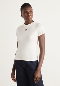 Trumpas rankovėmis kreminės spalvos marškinėliai, puošti spalvingu "Tommy Jeans" logotipu su tekstu "New York City", minkšta tekstūra ir prigludęs dizainas.