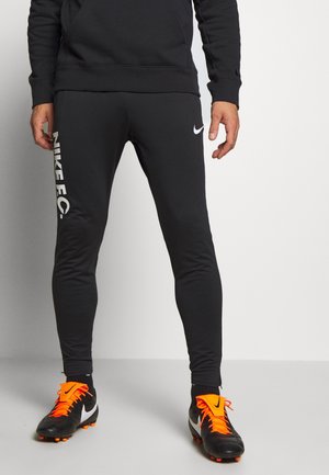 Mann trägt schwarze Nike FC Trainingshose und schwarze Fußballschuhe mit leuchtend orangefarbenen Schnürsenkeln und Akzenten, steht auf einem grauen Boden.