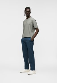 Chemise grise à manches courtes avec un logo discret, associée à un pantalon bleu et des chaussures blanches. La tenue présente une coupe décontractée et un design simple.