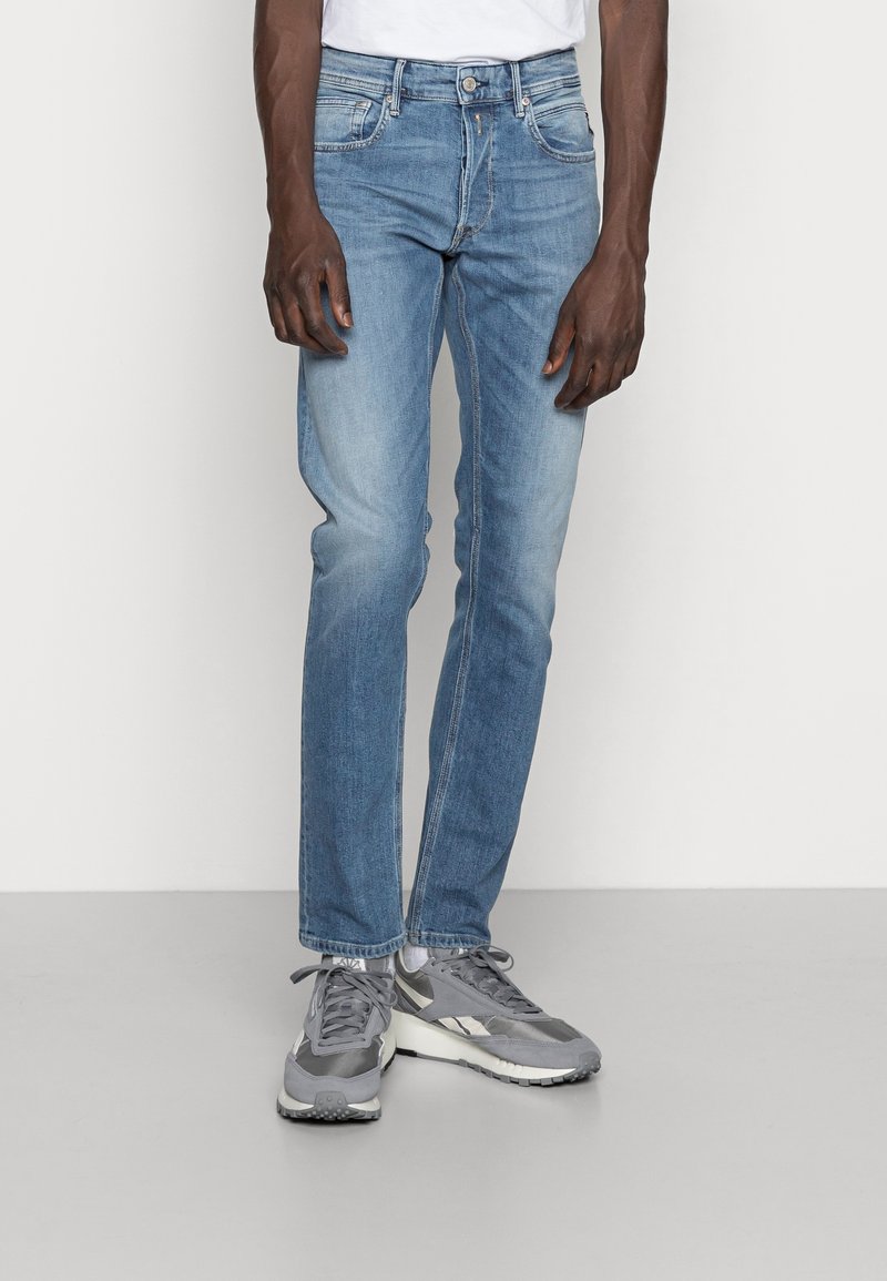 Jeans de mezclilla azul claro con un corte recto, que presentan un desvanecimiento sutil, cinco bolsillos y un cierre de botón. Se llevan con zapatillas deportivas grises.