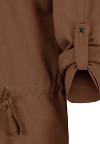 Manche en tissu marron avec un poignet roulé maintenu par une bride boutonnée et un détail cordon près de l'ourlet.