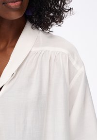 Biała, lekka bluzka z tkaniny o miękkiej fakturze, z wysmuklonym kołnierzykiem i marszczeniami na ramionach.