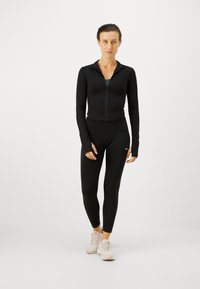 Svart zip-up träningsjacka i kombination med höghöga svarta leggings. Modellen har på sig ljusa sneakers. Slät, elastisk tyg.