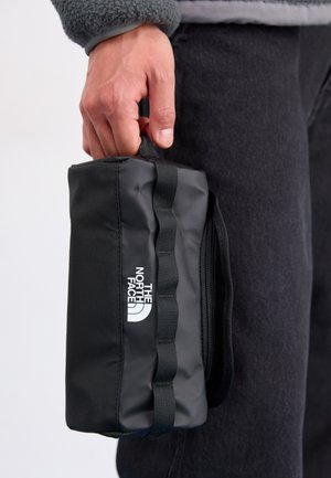 Trousse de toilette noire compacte avec une finition texturée, dotée d'une poignée supérieure, d'une fermeture éclair et d'un logo blanc à l'avant.