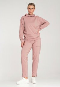 Dres z kapturem i spodniami w miękkim różowym materiale. Bluza ma luźny krój; spodnie mają dwie przednie kieszenie i rozcięcia u dołu.