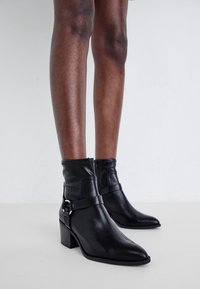 Black ankle boots made of leather, featuring a pointed toe, a low stacked heel, a zip closure, and a decorative strap with a metal accent.
