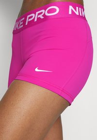 Zbliżenie na osobę ubraną w jasnoróżowe spodenki sportowe Nike Pro z białym logo Nike na udzie oraz markowym pasem.
