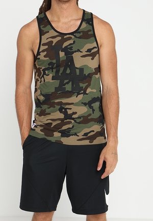 Mężczyzna ubrany w maskującą koszulkę na ramiączkach z czarnym logo "LA" oraz czarne sportowe szorty z białym znakiem Nike, stojący z rozluźnionymi rękami.