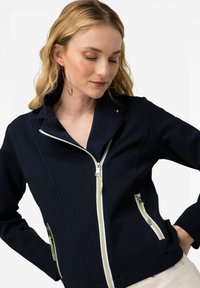 Chaqueta de navy con cremallera y cuello con muesca, acentos amarillos y dos bolsillos con cremallera. Hecha de una tela texturizada y elástica.