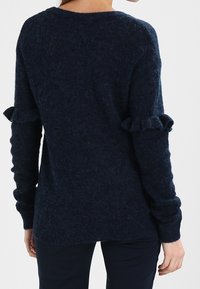 Marineblauwe gebreide trui met een textuurafwerking, lange mouwen met ruches en een ronde halslijn. Achteraan is een ontspannen pasvorm te zien.