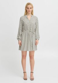 Rochie cu mânecă scurtă dintr-un material ușor, cu un decolteu cu nasturi, talie cu șnur și un model cu buline întunecate pe un fundal crem.